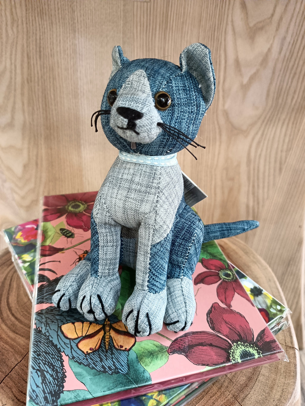 Estatueta de gato em tecido cinzento e azul sobre livros florais e madeira