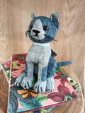 Estatueta de gato em tecido cinzento e azul sobre livros florais e madeira
