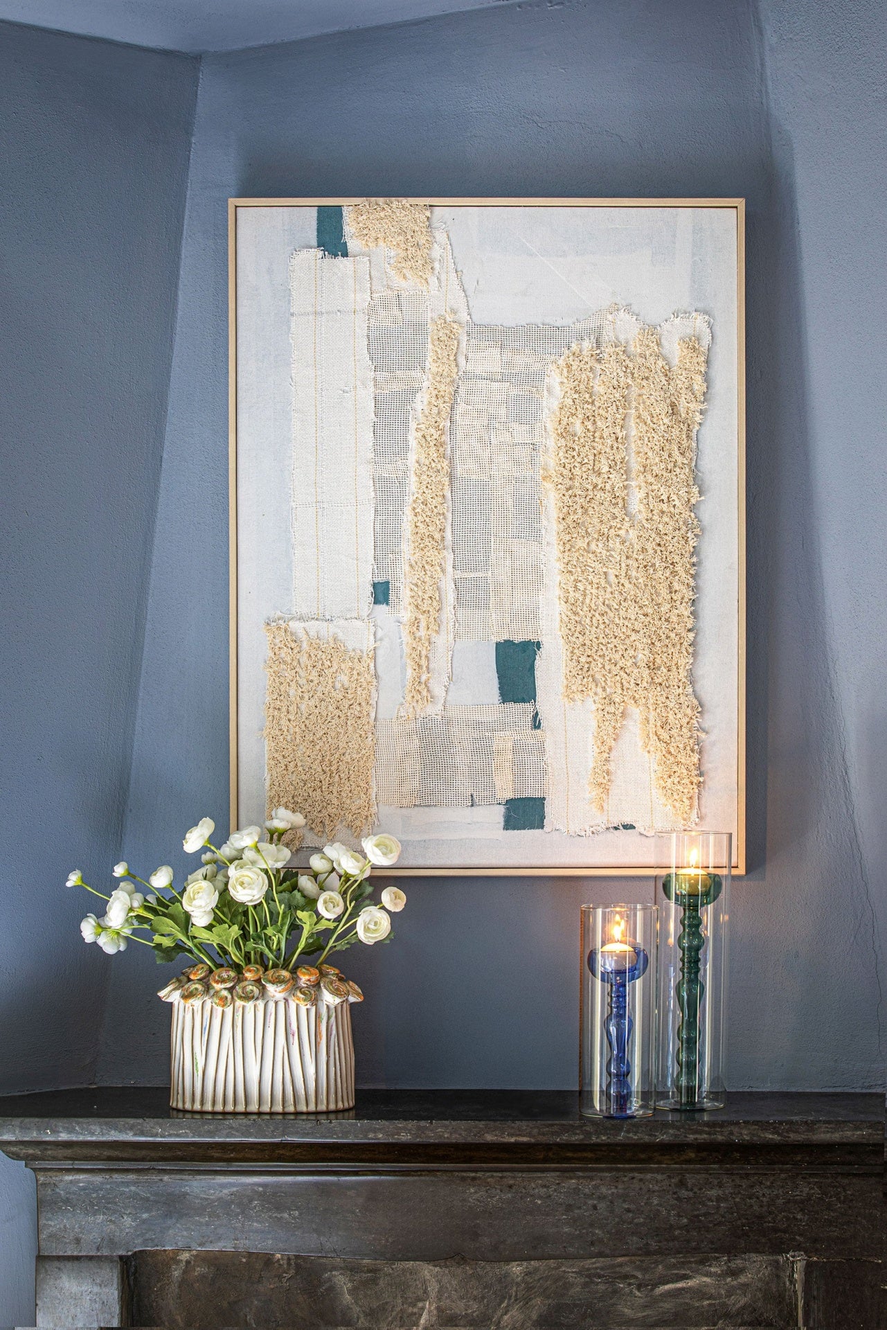 Quadro texturizado de tecidos naturais em tons creme e azul na parede cinzenta, com jarro de flores brancas e castiçais de vidro sobre lareira.