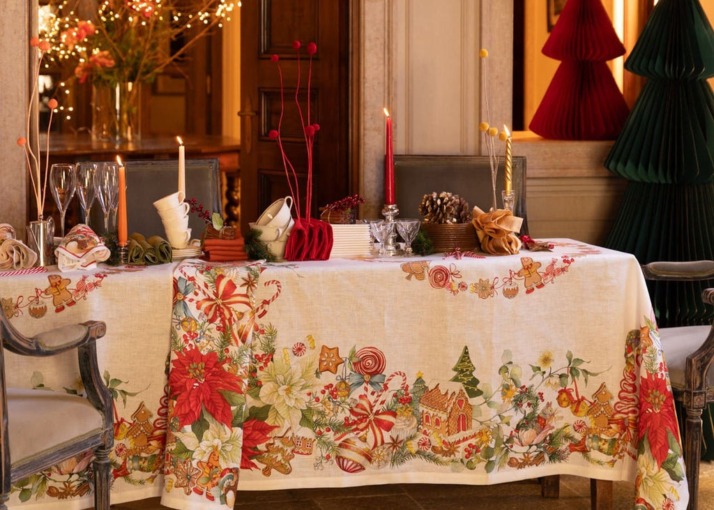 Toalha de mesa natalícia com padrão de flores, doces e bolachas, mesa posta com velas, copos, chávenas e árvores de papel decorativas ao fundo.
