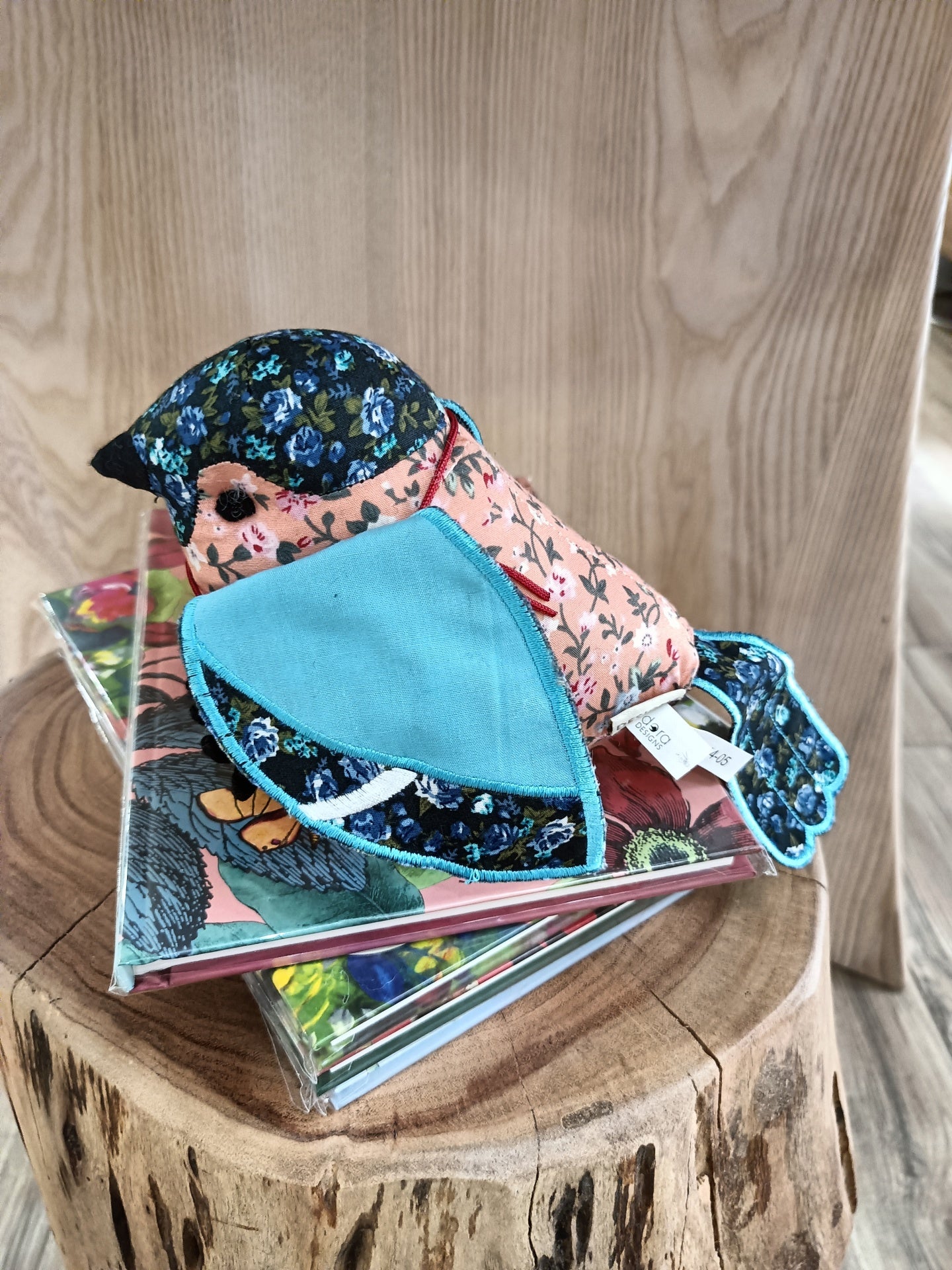 Boneco de tecido em forma de pássaro com padrão floral azul e rosa, sobre livros e tronco de madeira