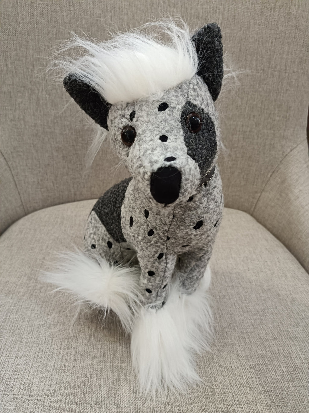 Boneco cão de peluche cinzento e negro com pelo branco artificial sentado num sofá cinzento.