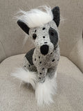 Boneco cão de peluche cinzento e negro com pelo branco artificial sentado num sofá cinzento.