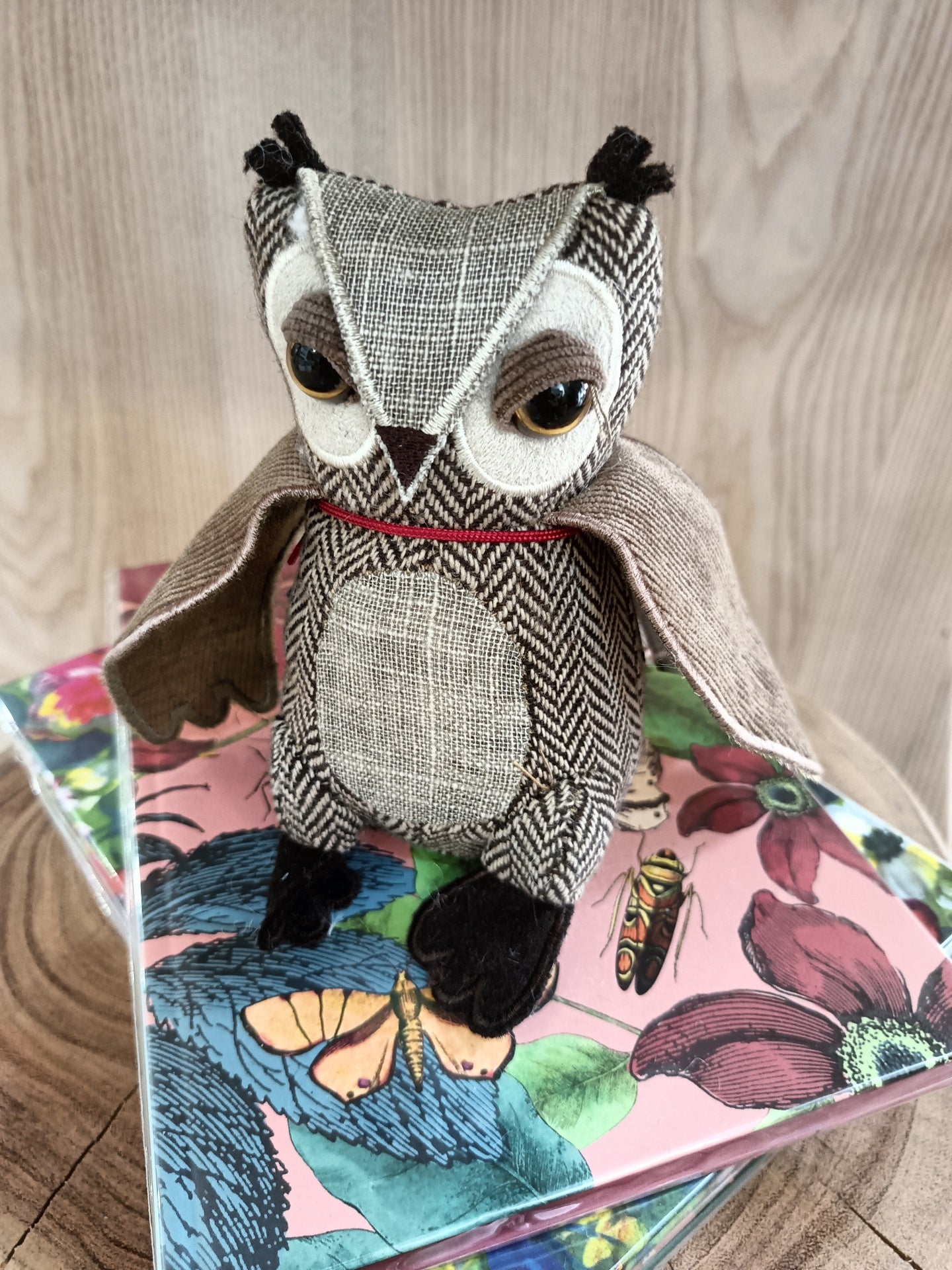 Boneco de coruja de tecido cinza e bege com olhos grandes sobre caixas coloridas com estampas de flores e borboletas