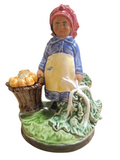 Estatueta cerâmica de uma mulher com lenço rosa, vestido azul, avental amarelo, com cesta de abóboras e vegetais verdes.