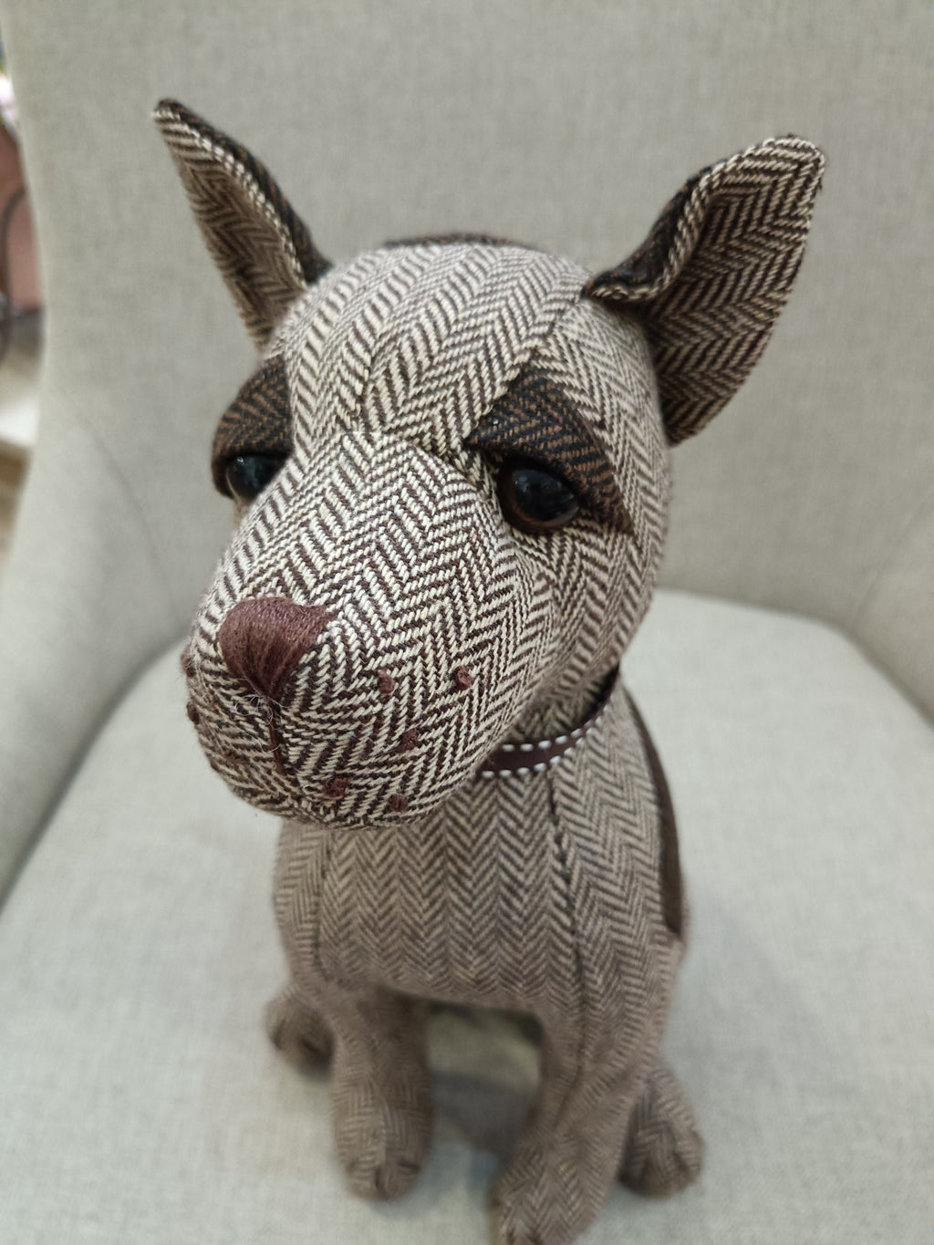 Boneco decorativo de cão em tecido espinha de peixe castanho e bege sentado em cadeira cinza.