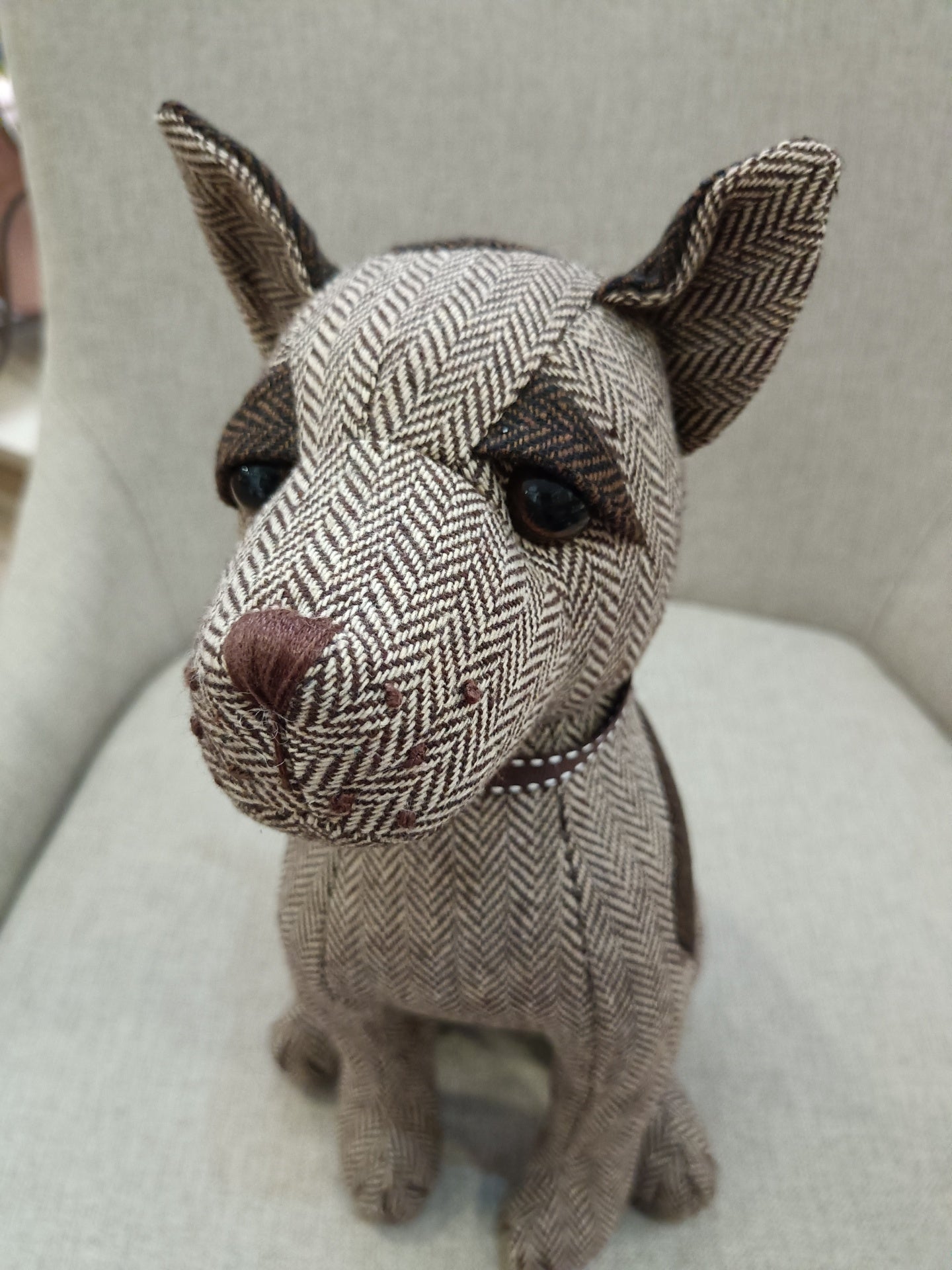 Boneco decorativo de cão em tecido espinha de peixe castanho e bege sentado em cadeira cinza.
