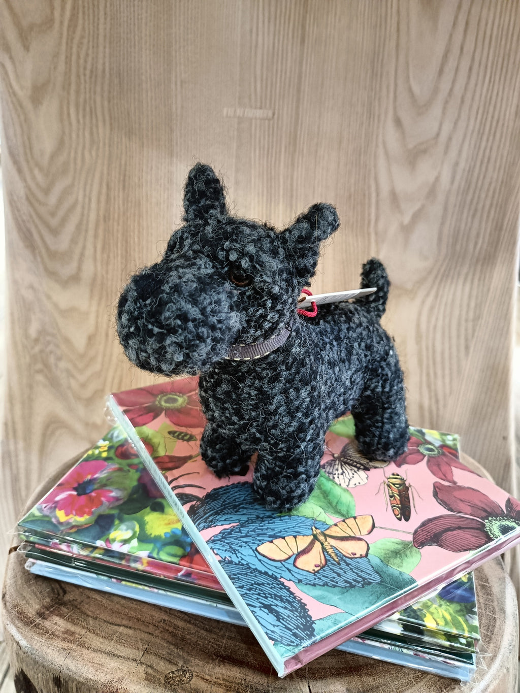 Miniatura de cão de pelúcia cinzento escuro com coleira roxa junto a cadernos coloridos com flores e borboletas sobre banco de madeira