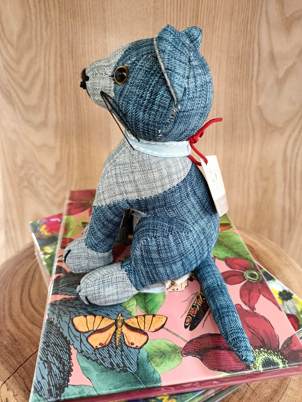 Urso de peluche azul e cinzento em tecido riscado com etiqueta e laço vermelho sentado sobre caixa floral colorida