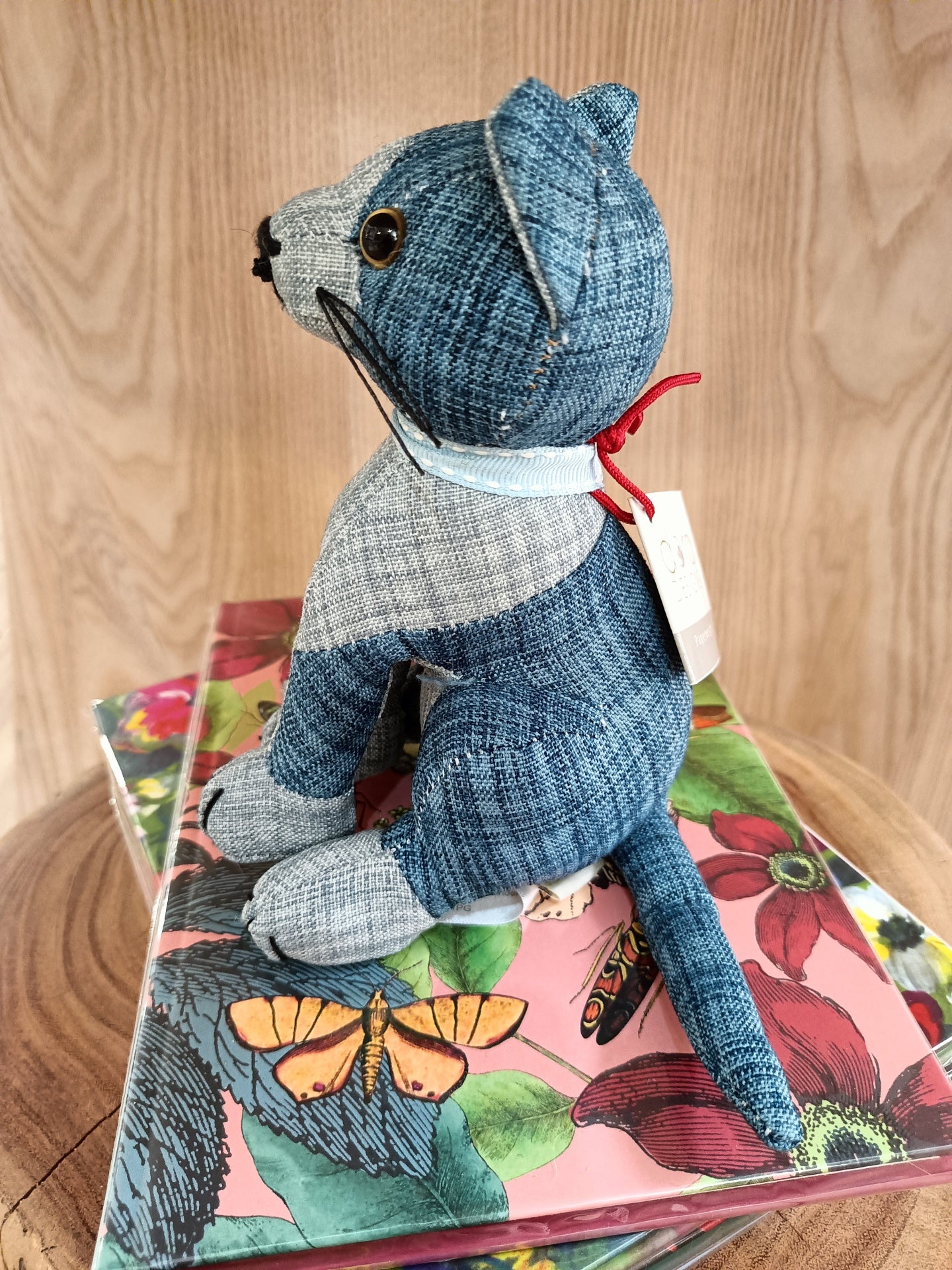 Urso de peluche azul e cinzento em tecido riscado com etiqueta e laço vermelho sentado sobre caixa floral colorida