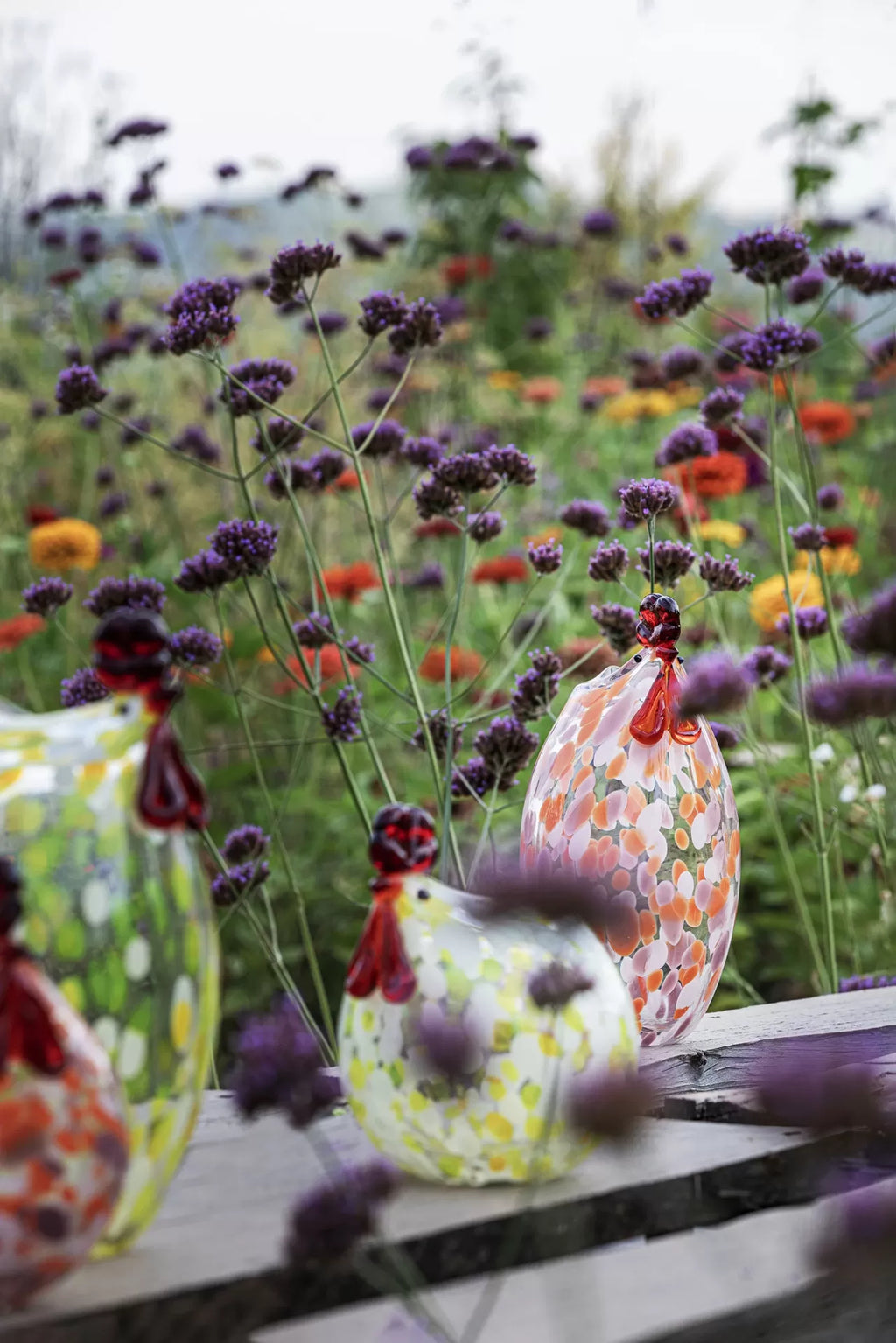 Objetos decorativos em vidro em forma de galinhas coloridas sobre mesa de madeira com flores violetas atrás.