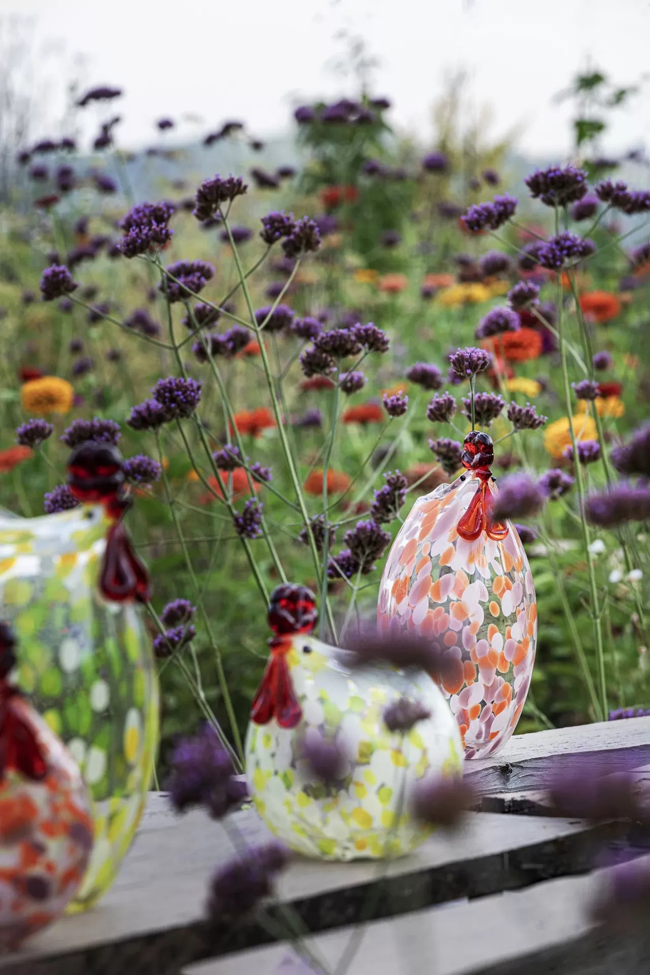 Objetos decorativos em vidro em forma de galinhas coloridas sobre mesa de madeira com flores violetas atrás.