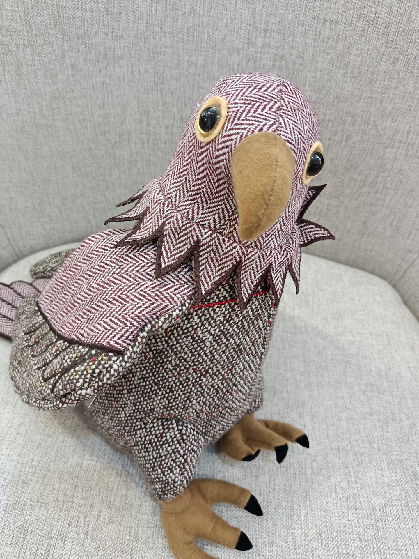 Pássaro decorativo em tecido com padrões vermelhos, brancos e cinzentos, com bico castanho e olhos grandes, no sofá cinzento.