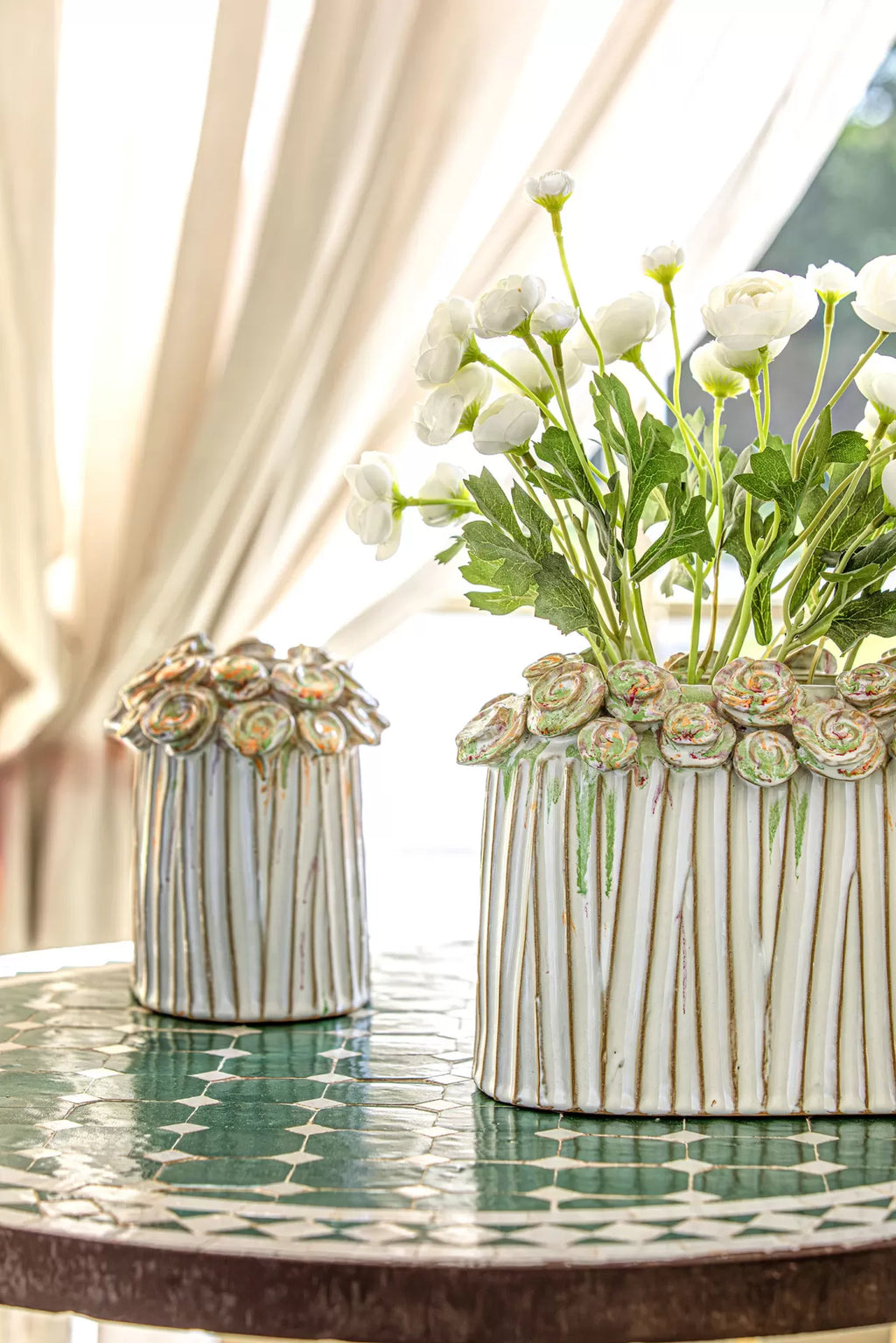 Dois vasos cerâmicos decorativos com rosas em relevo, um com flores artificiais brancas, sobre mesa com azulejos verdes e bege.