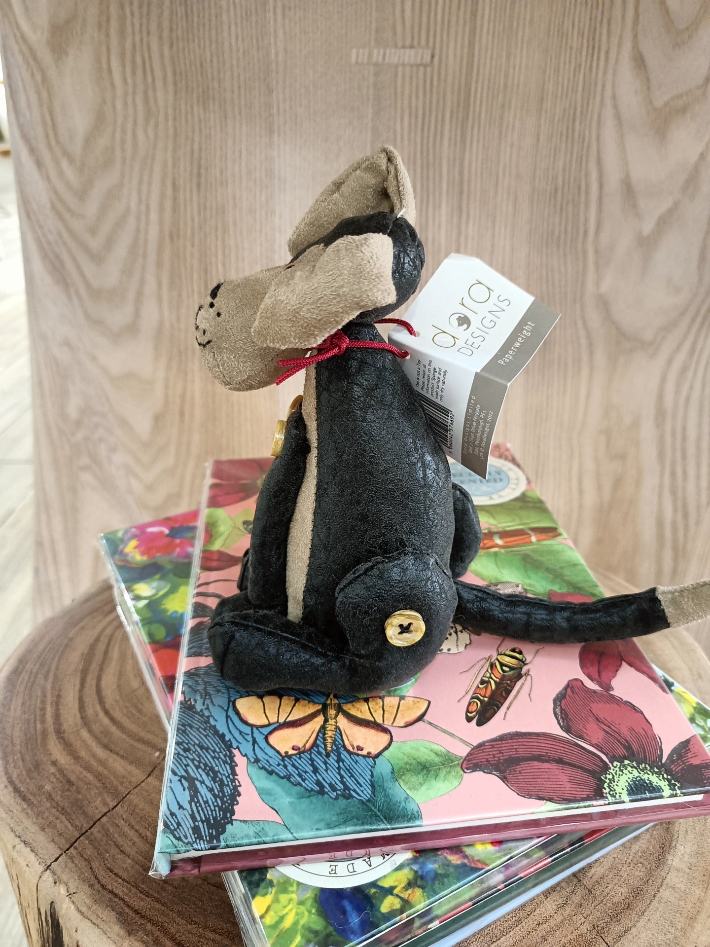 Ratinho de pelúcia preto e bege com laço vermelho e etiqueta, sobre livros florais numa mesa de madeira