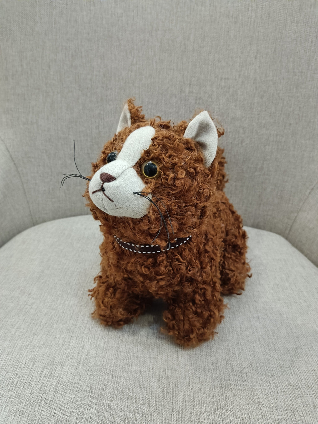 Pelúcia de gato castanho com detalhes brancos e olhos grandes sentada num sofá cinzento