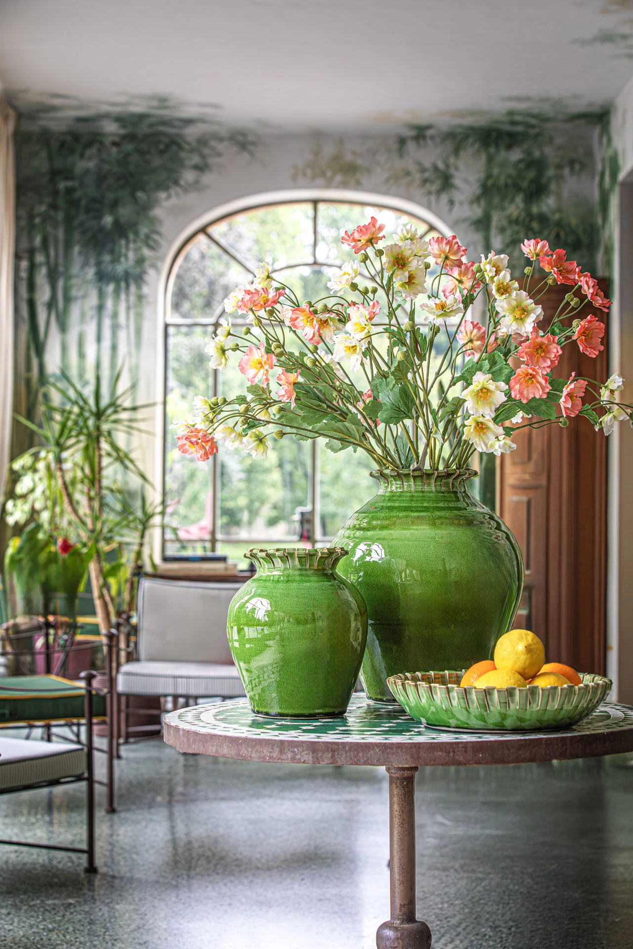 Vasos verdes com flores artificiais sobre mesa redonda com tampo em mosaico num interior com janela arqueada.