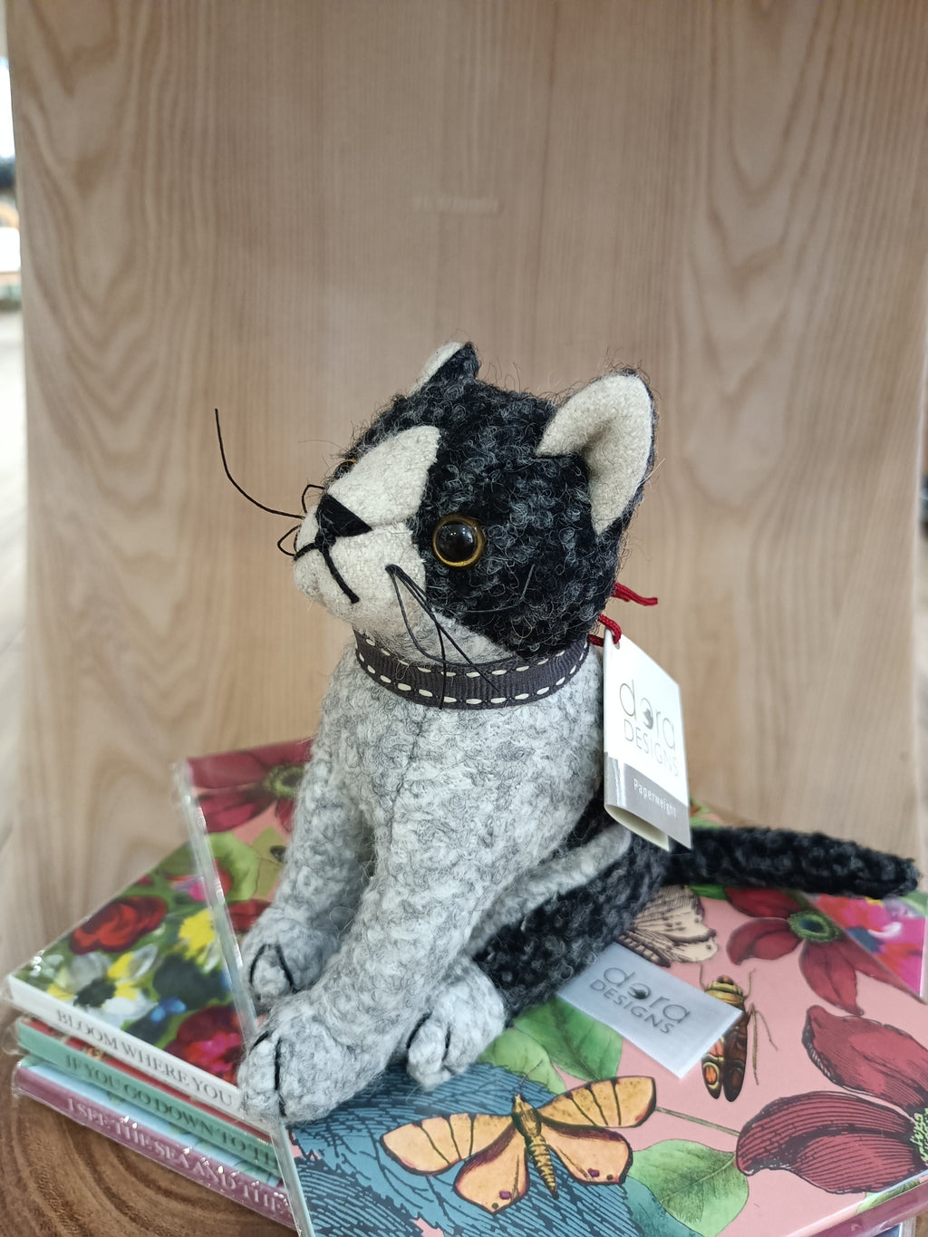 Boneco decorativo de gato em tecido cinzento e preto sobre livros coloridos com capa ilustrada.