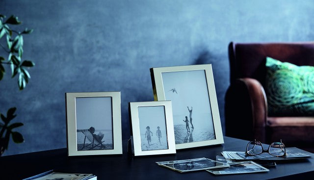 Três molduras de fotografias em tons sépia numa mesa com óculos e revistas, com sofá castanho ao fundo