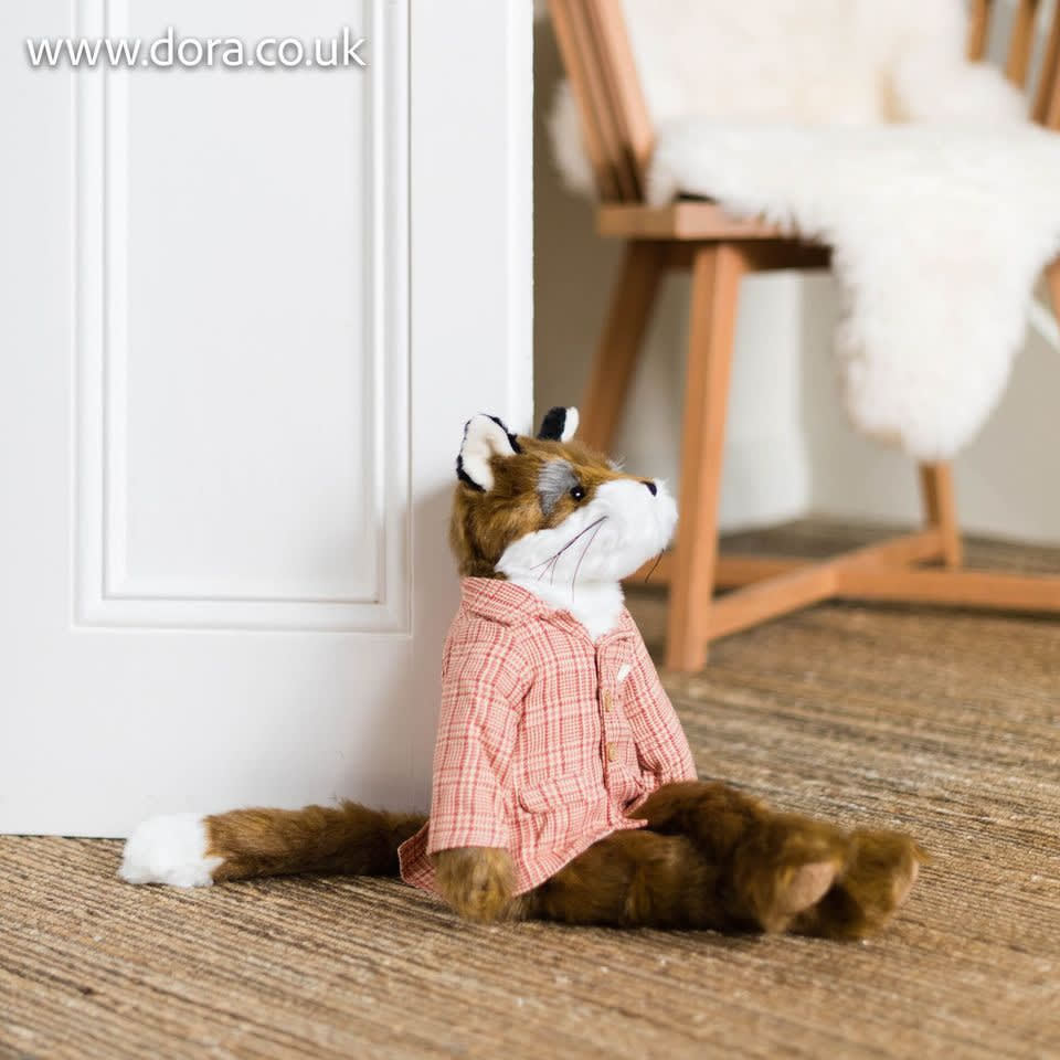 Boneco de raposa de pelúcia vestido com camisa xadrez rosa, sentado no chão junto a uma porta e cadeira