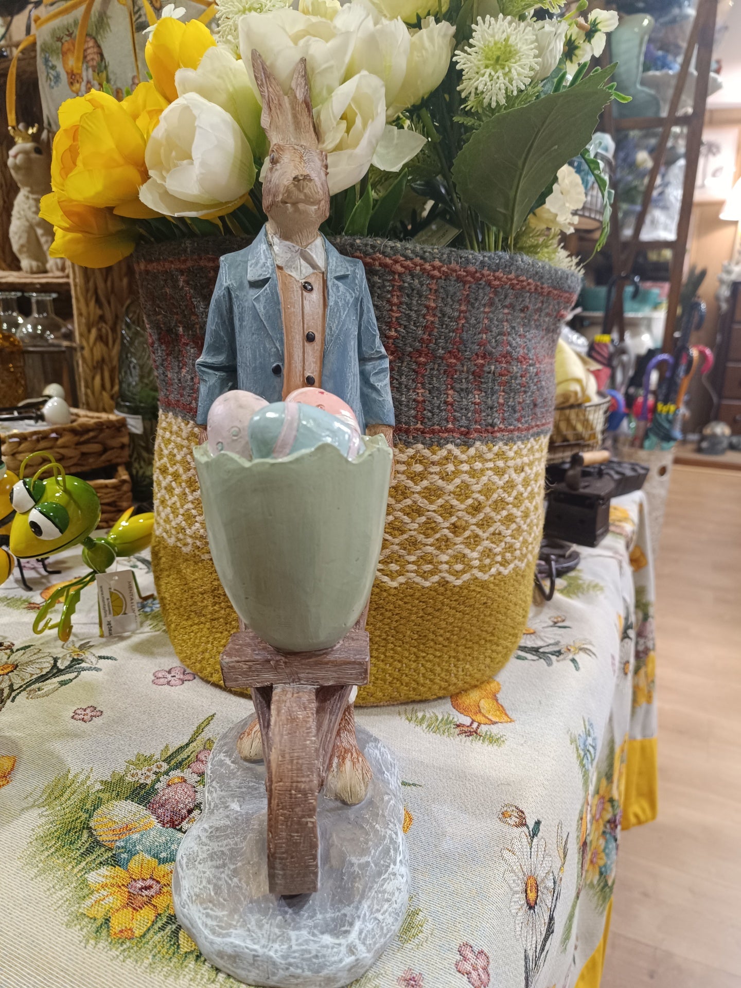 Figura decorativa de coelho a empurrar carrinho com ovos junto a vaso com flores sobre mesa com toalha floral