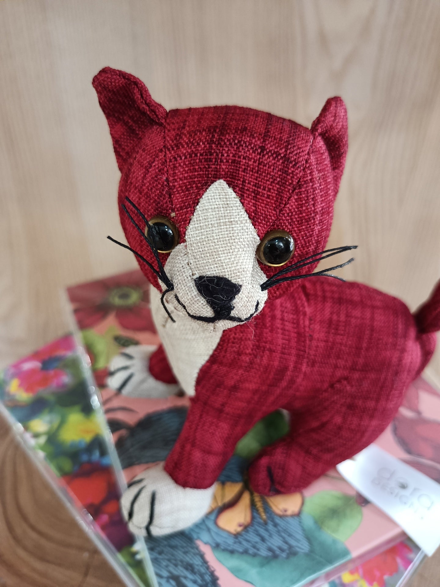 Gato de peluche vermelho e creme em tecido junto a caixa floral