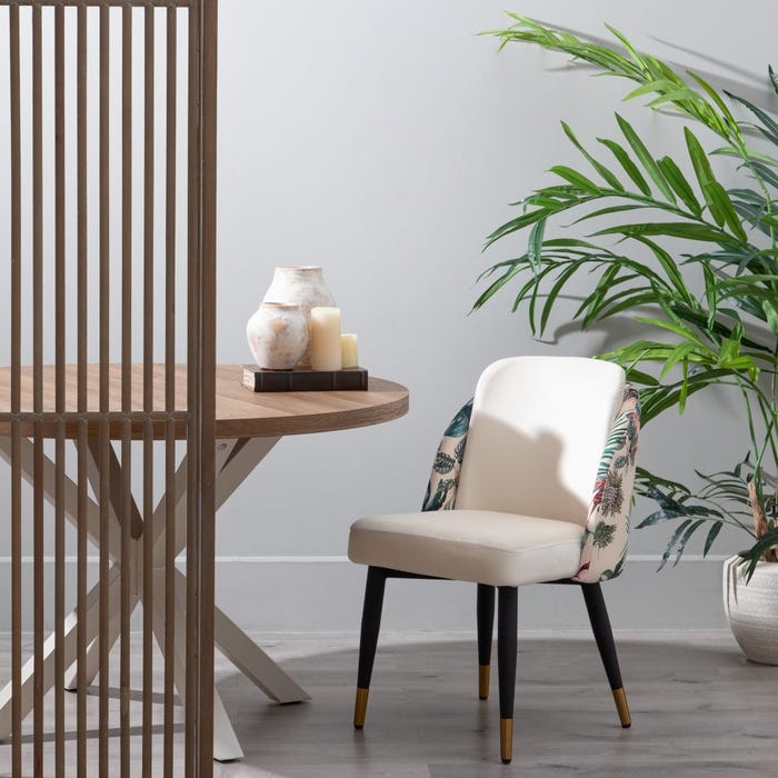 Cadeira estofada creme com padrão floral, mesa de madeira redonda, vela, vaso decorativo e planta verde
