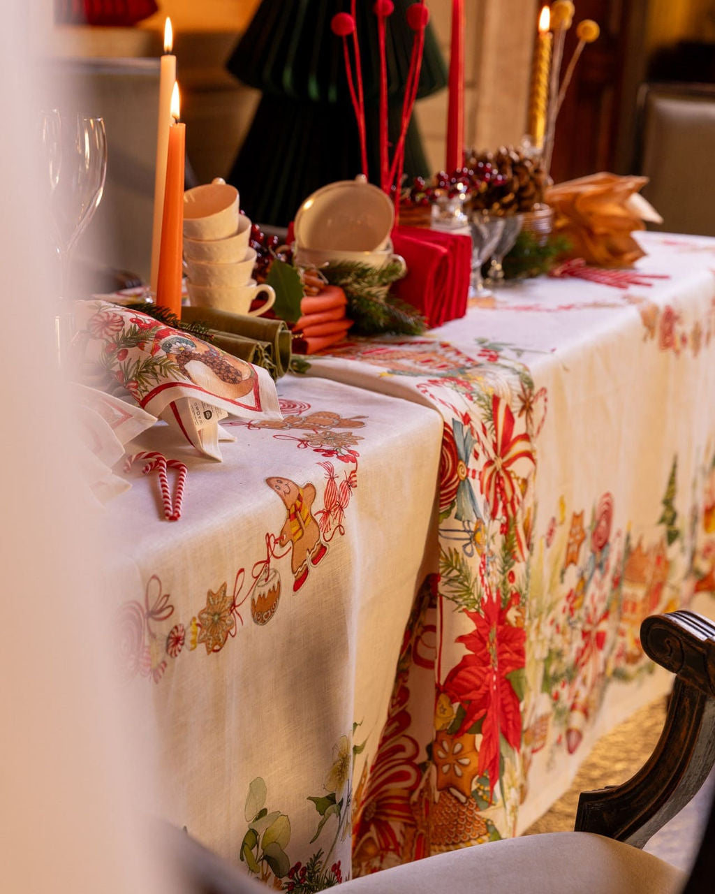 Mesa decorada para Natal com toalha branca e estampas natalinas, velas acesas, chávenas e guardanapos