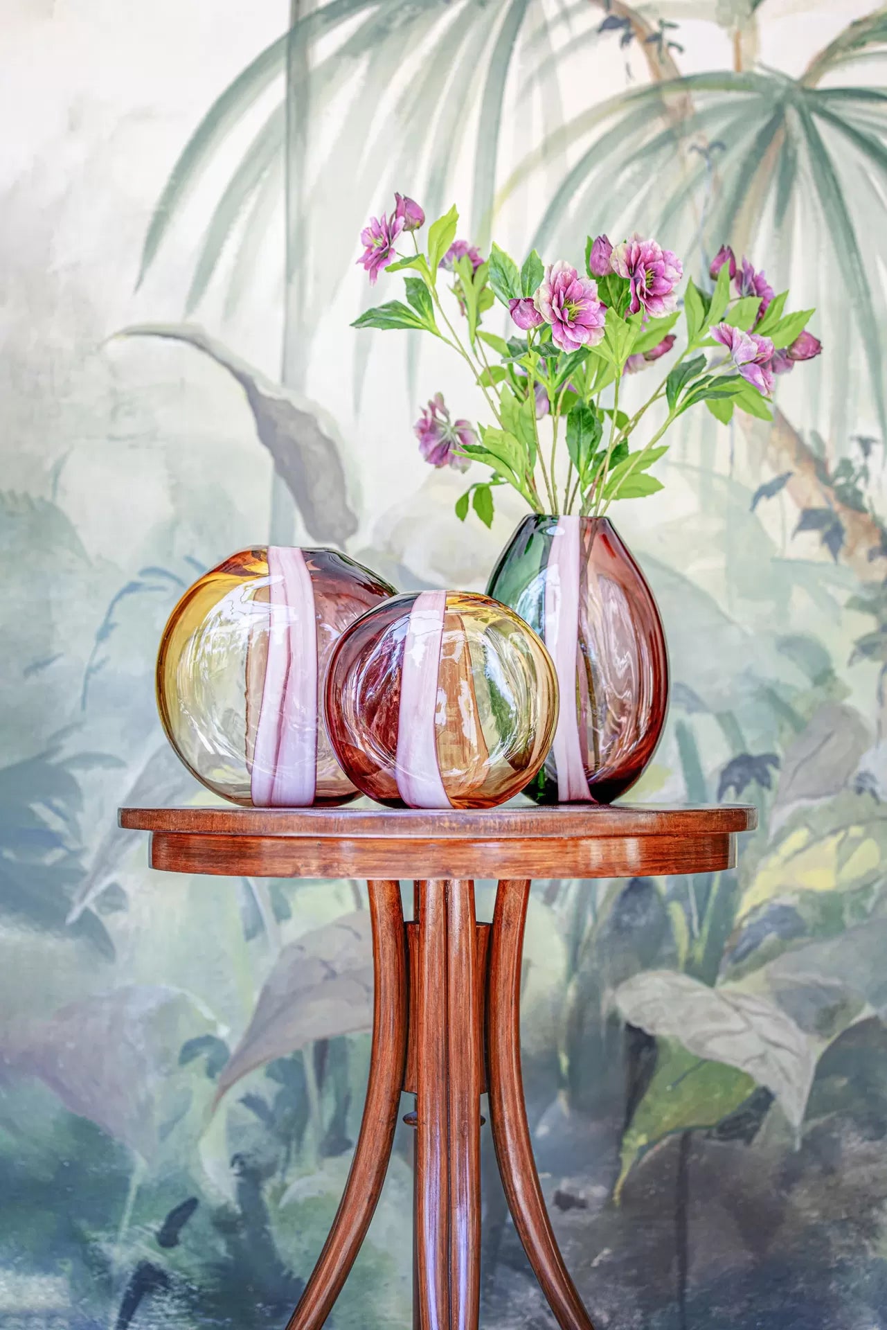 Três vasos coloridos em vidro com flores sobre mesa de madeira e fundo com pintura botânica.