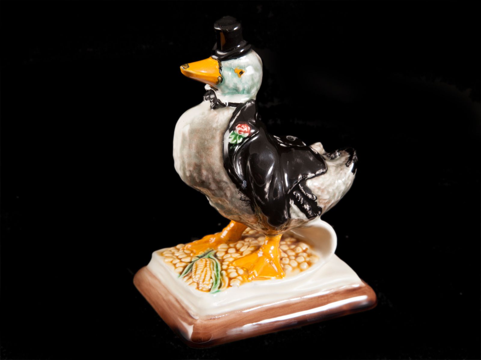 Figuinha de cerâmica pato com vestuário preto e base decorada.