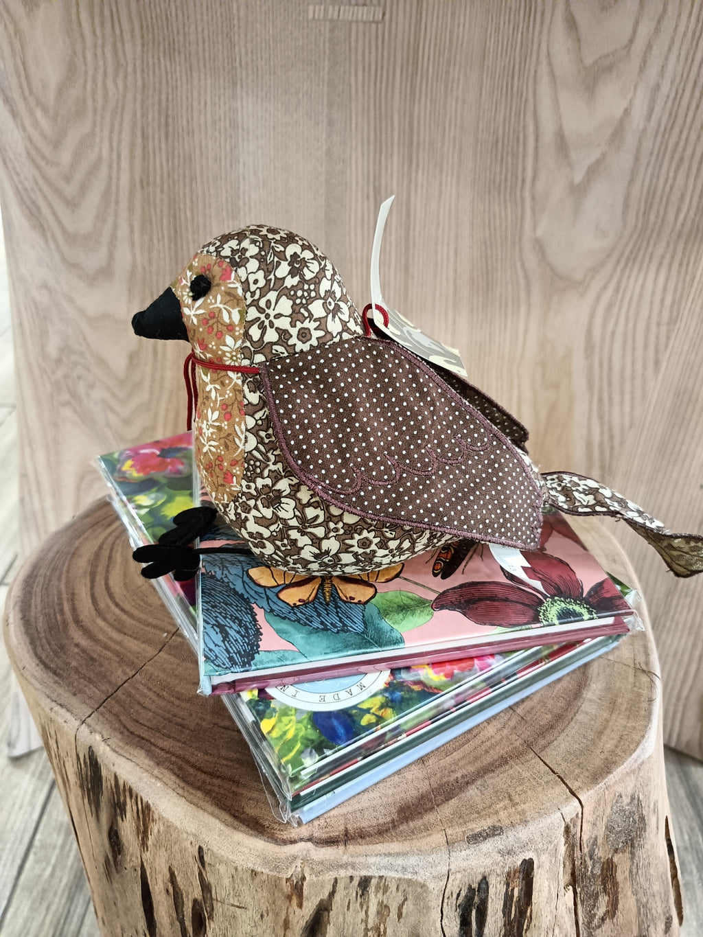 Pássaro decorativo de tecido com padrão floral castanho e beige sobre cadernos florais e tronco de madeira