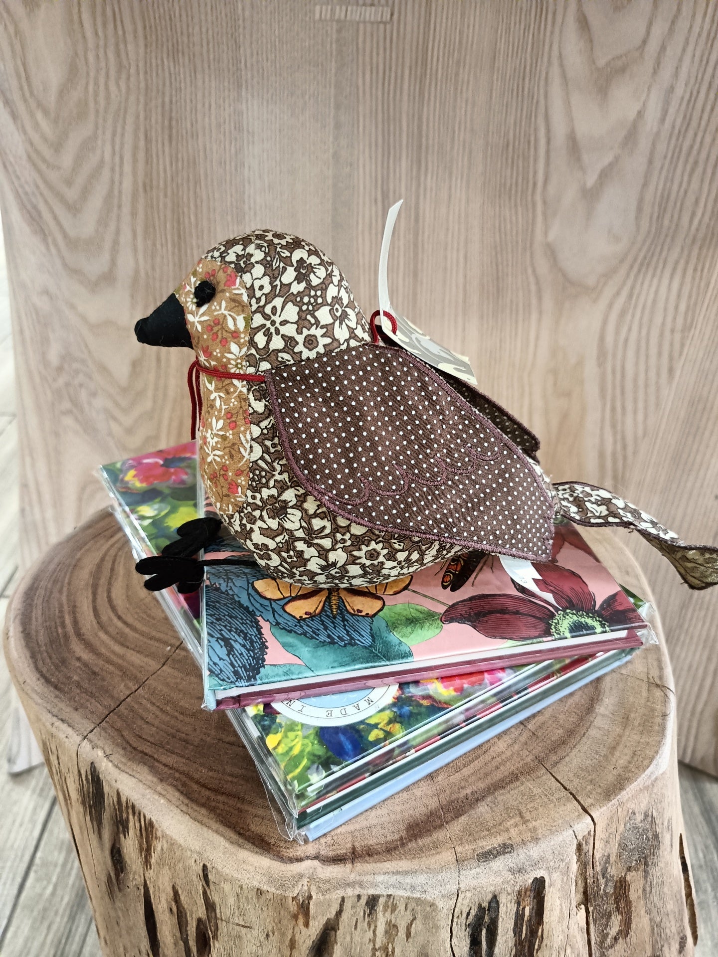 Pássaro decorativo de tecido com padrão floral castanho e beige sobre cadernos florais e tronco de madeira