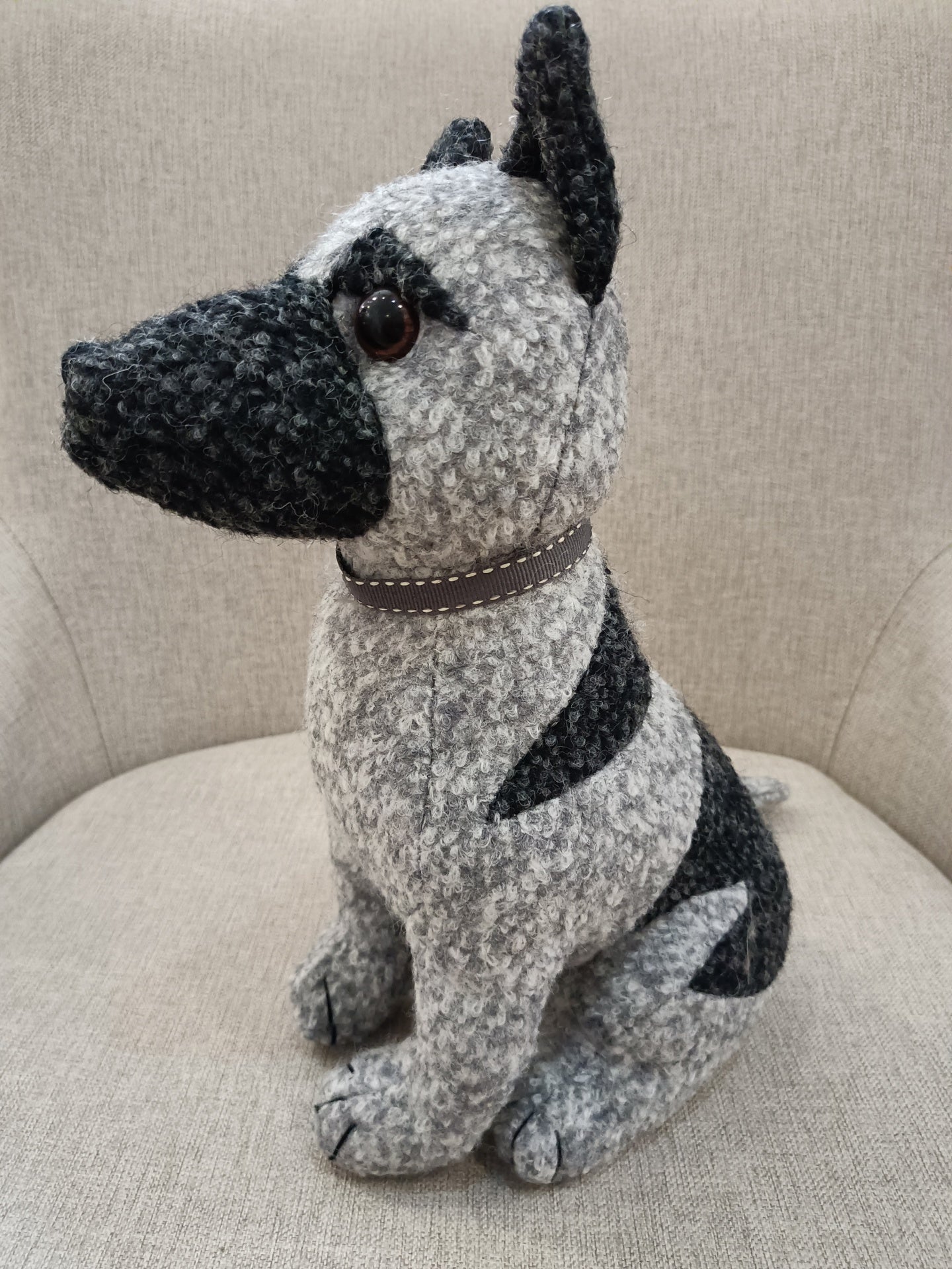 Pelúcia em forma de cão cinzento com manchas negras e olhos castanhos sentada num sofá cinzento claro