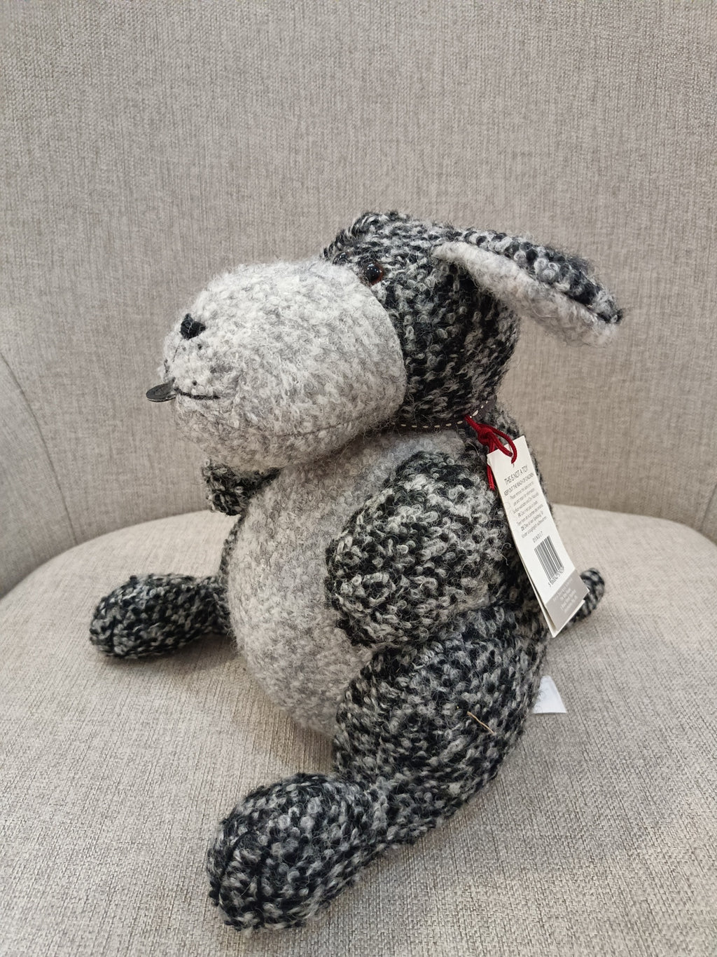 Ursinho de pelúcia cinzento e preto sentado num sofá cinzento com etiqueta pendurada