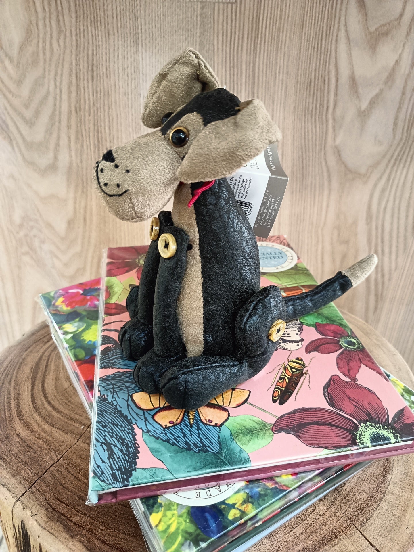 Boneco cachorro de tecido preto e bege com botões, sobre caixas decorativas florais numa mesa de madeira
