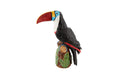 Estatueta decorativa de tucano colorido sobre tronco com folhas verdes.