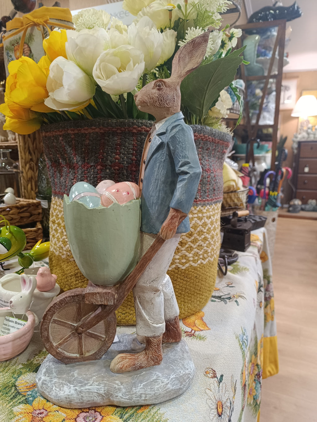 Estatueta de coelho a empurrar carrinho com ovos coloridos, vaso de flores e toalha estampada