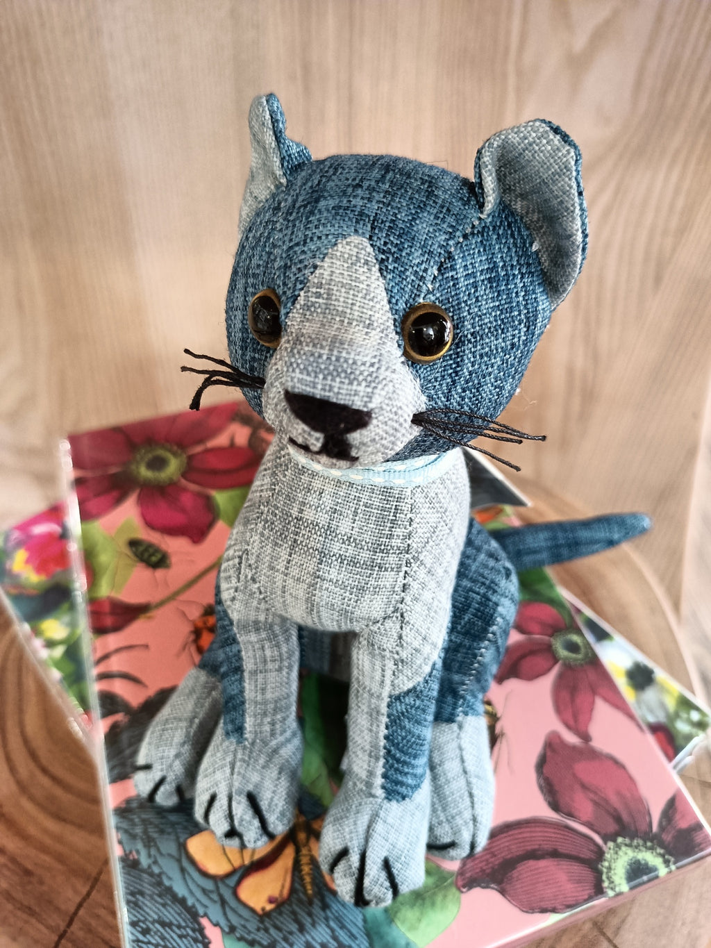 Boneco de gato tecido azul e cinza com olhos castanhos sentado em caixa floral