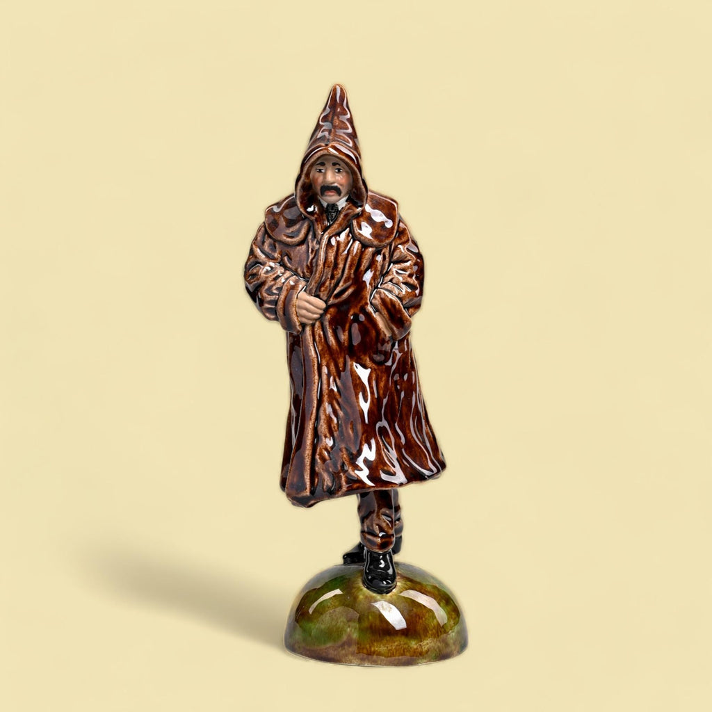 Estatueta cerâmica homem casaco marrom capuz base verde.