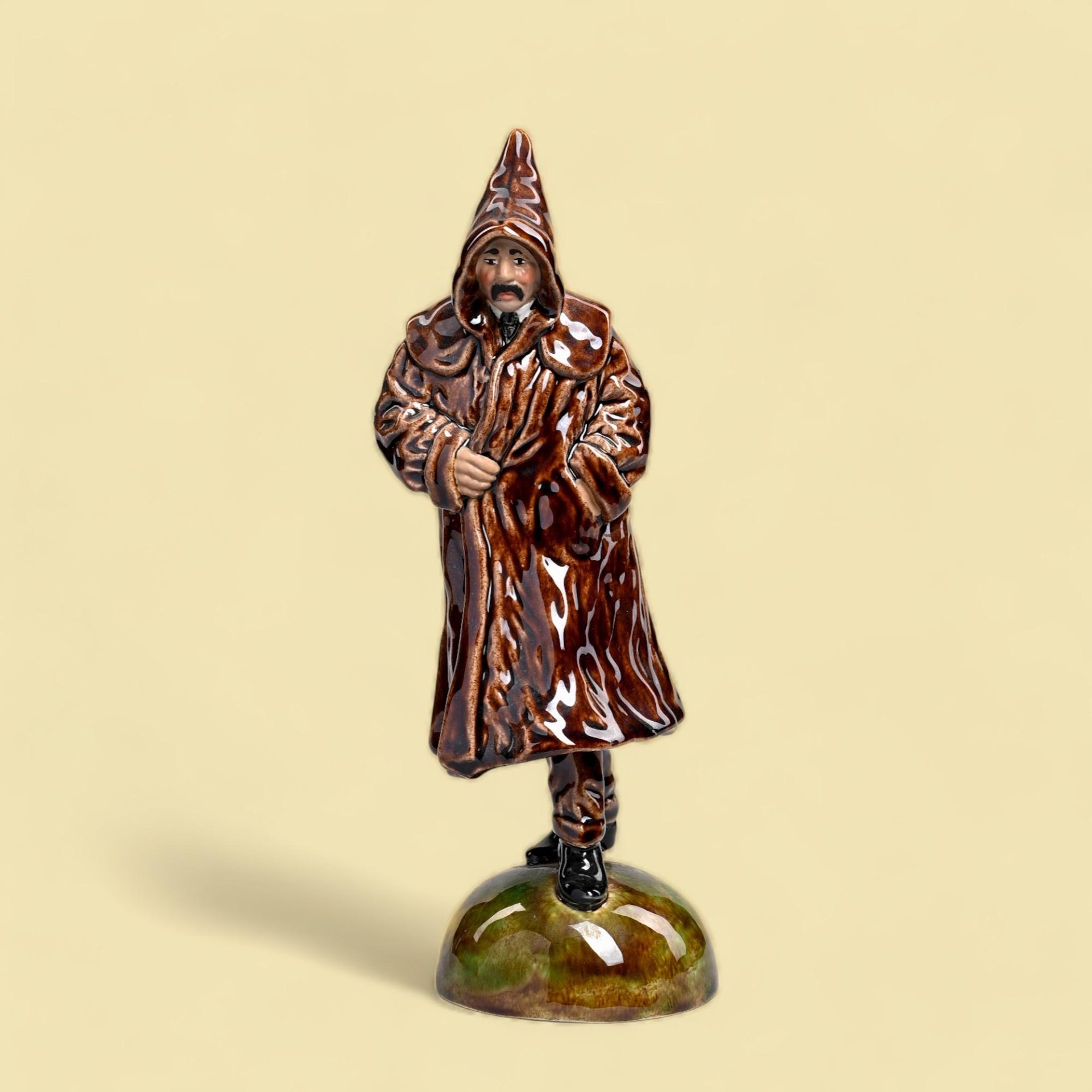 Estatueta cerâmica homem casaco marrom capuz base verde.